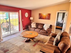 A living room with a brown leather sofa, a wooden coffee table, and a patterned rug. A red wall and a door to a bedroom are visible.