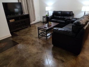 A living room with a dark brown leather sofa, a coffee table, a TV on a stand, and two lamps on side tables. - Chateau 13, spacious townhouse in Tickfaw, LA (Tickfaw)
