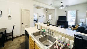 A compact kitchen with a double sink, a dining area with a table and chairs, a ceiling fan, and a view of the living room with a sofa and a TV.