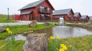 Una cabaña roja de madera con balcón, un pequeño estanque con cascada y narcisos en primer plano.