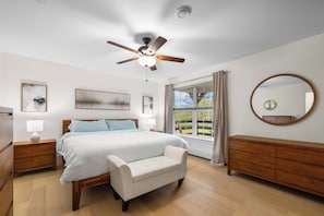 A bedroom with a bed, a ceiling fan, a bedside table with a lamp, a dresser, a window with curtains, and a round mirror on the wall.