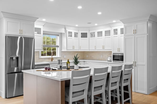 A modern kitchen with white cabinetry, a stainless steel refrigerator, a central island with bar stools, and a window providing natural light.