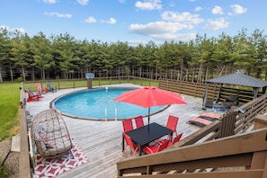 A wooden deck with a swimming pool, red chairs, a table, and a gazebo.