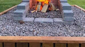Un foyer avec des bûches et un bord en pierre, entouré de gravier et d’une terrasse en bois.