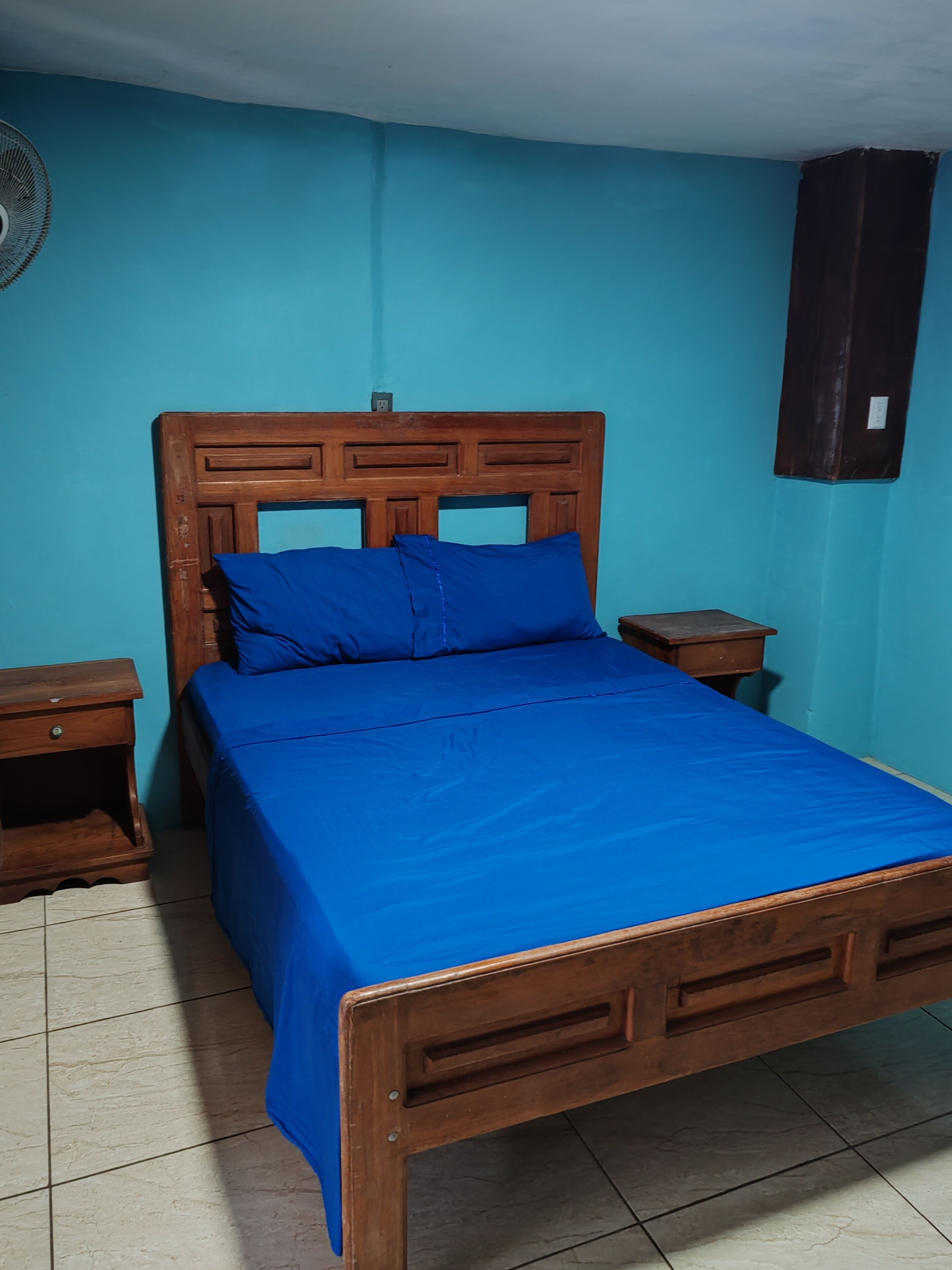 Ein Zimmer mit einem Einzelbett, einem Holzkopfstück, einer blauen Bettdecke, zwei Nachttischen und einem an der Wand befestigten Ventilator.