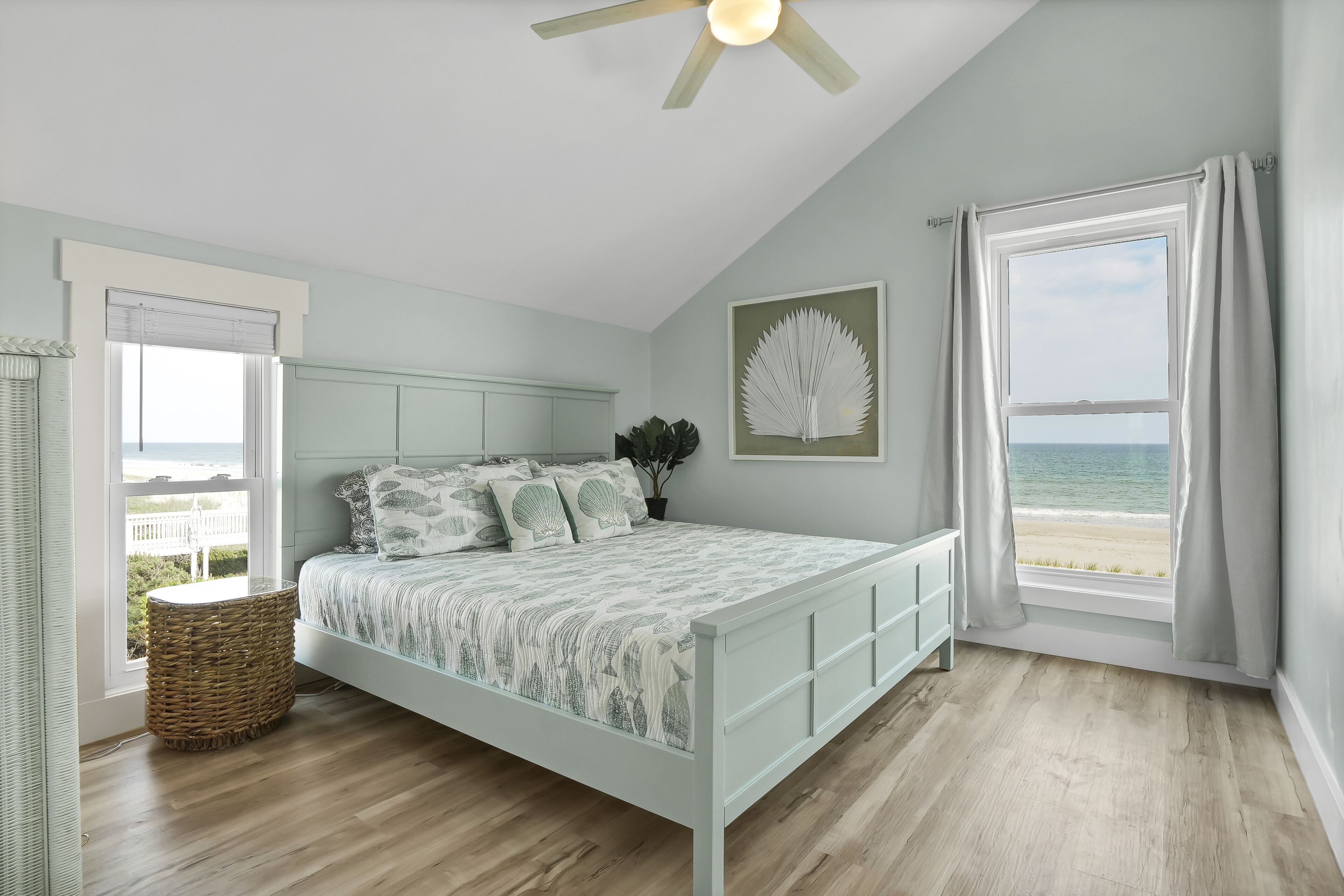 Ein Schlafzimmer mit Bett, Fenster mit Meerblick, Deckenventilator und Wandbild.