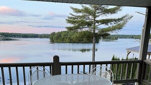 Une terrasse extérieure couverte, dotée d’une table et de chaises en métal rondes, qui offre une vue sur un lac paisible, bordé d’arbres et d’une rive lointaine.
