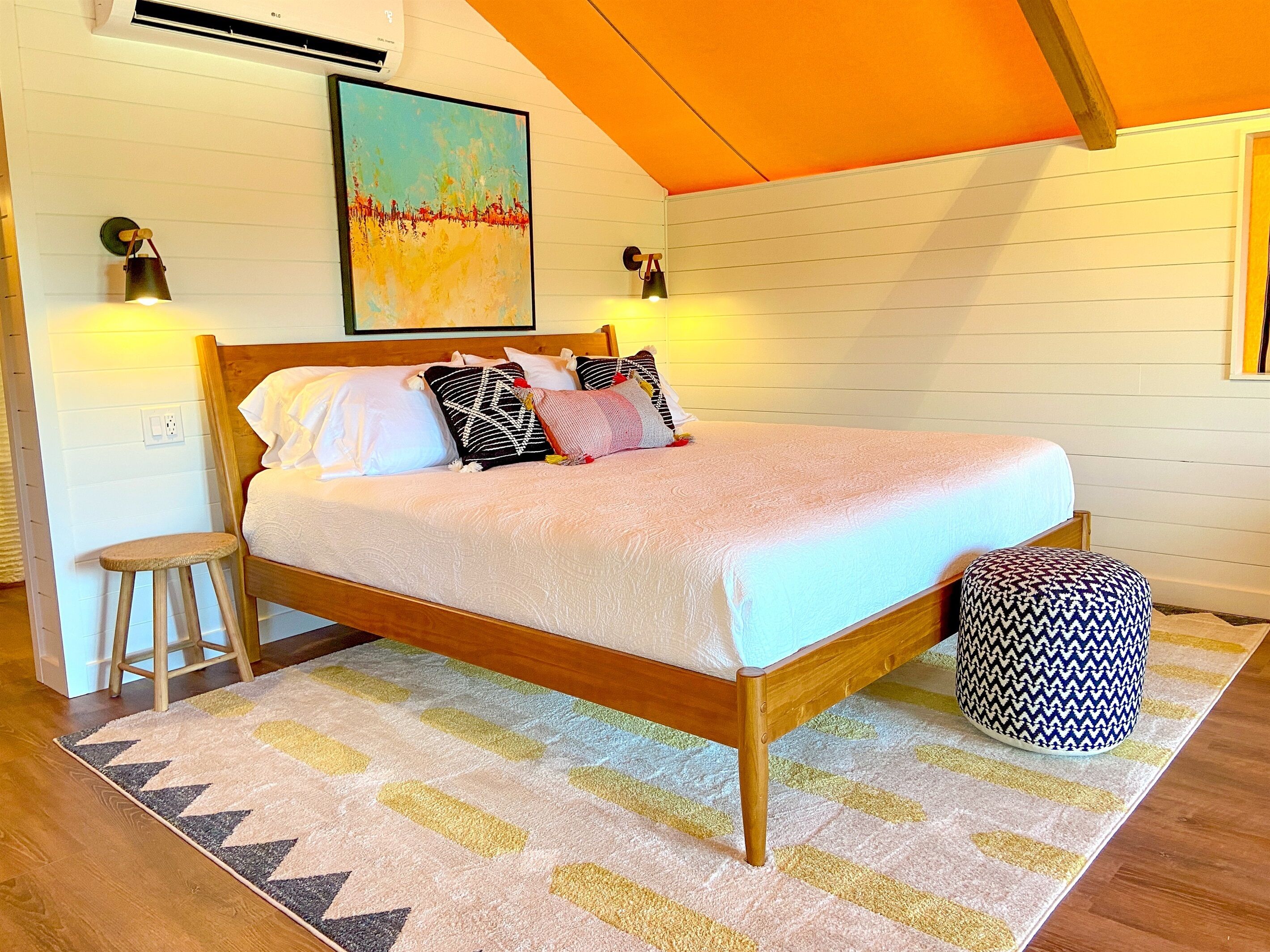 Un dormitorio con una cama de madera, un tapete colorido, un taburete pequeño y un otomano estampado.