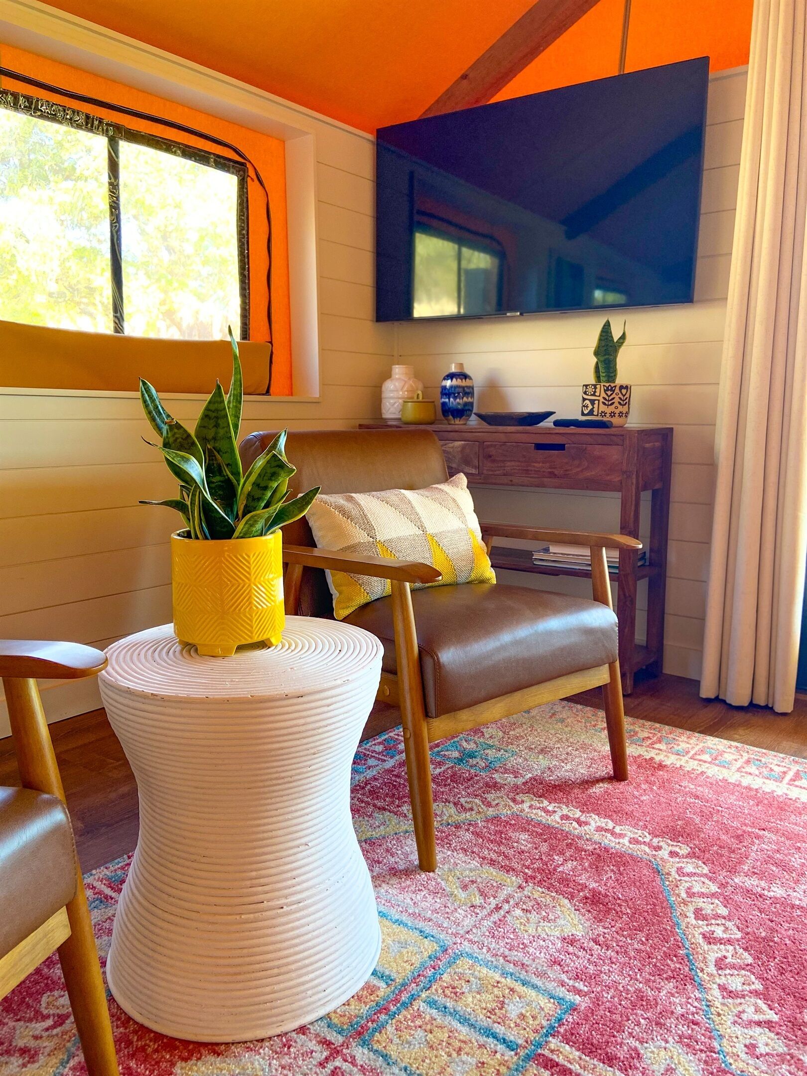 A cozy living room with a wooden floor, a round white table with a plant, a brown leather chair, a TV mounted on the wall, and a window with curtains.