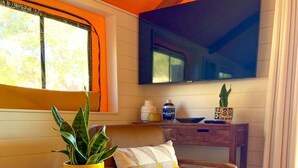 Un salon chaleureux avec un plancher en bois, une table ronde blanche agrémentée d’une plante, un fauteuil en cuir marron, un téléviseur fixé au mur et une fenêtre avec des rideaux.