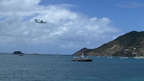 Une scène côtière avec des bateaux, un voilier et un avion à réaction survolant l’eau.