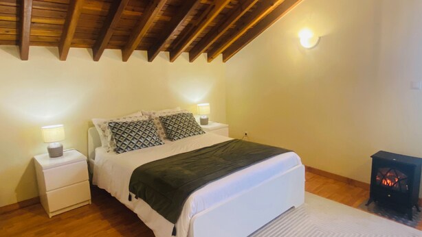 Une chambre avec un plafond en bois, un lit agrémenté d’oreillers à motifs, des lampes de chevet et un poêle à bois.