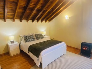 Un dormitorio con techo de madera, cama con almohadas estampadas, lámparas de noche y un estufa de leña.