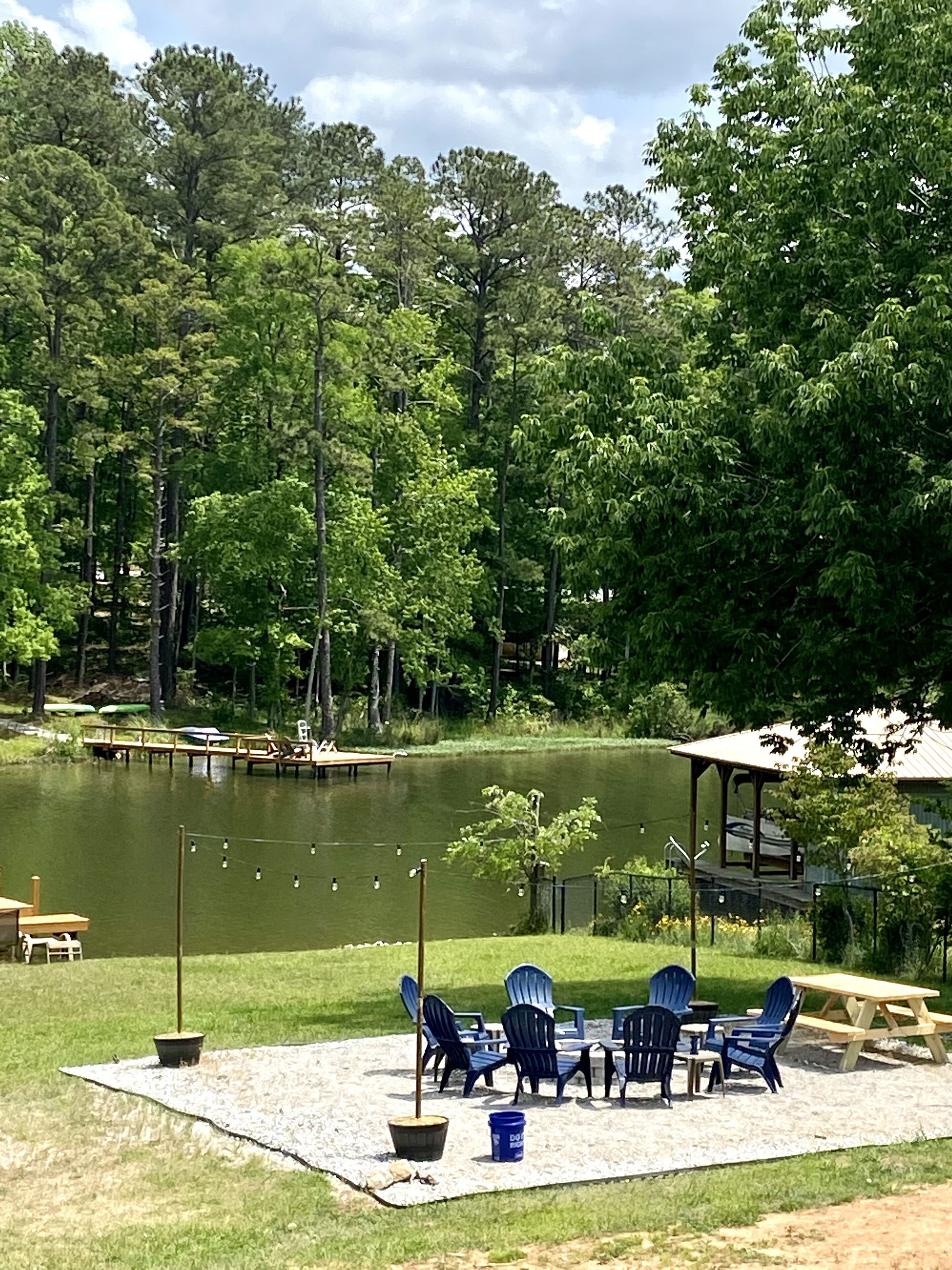 Un muelle junto al lago con sillas Adirondack y una mesa de picnic, rodeado de árboles frondosos y un embarcadero.