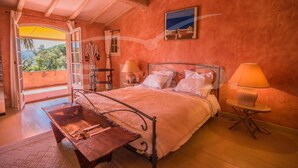 A bedroom with a bed, bedside lamps, a wooden table, and a view through an open door. - Villa with pool and sea view in Grimaud (GRIMAUD)