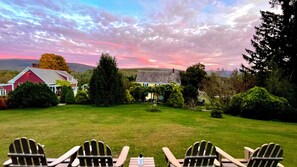 A grassy outdoor area with several Adirondack chairs, a picnic table, and a view of a house with a red roof and white trim.