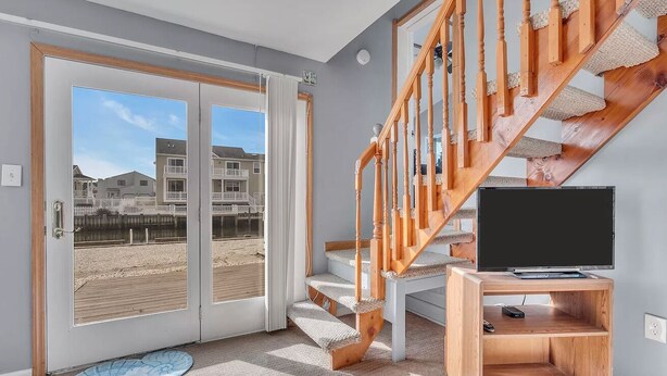 A modern interior with a wooden staircase, a television on a wooden stand, and a large glass door leading to a balcony with a view of buildings and a beach. - Lagoon-front home just minutes from the beach! (Stafford Township)