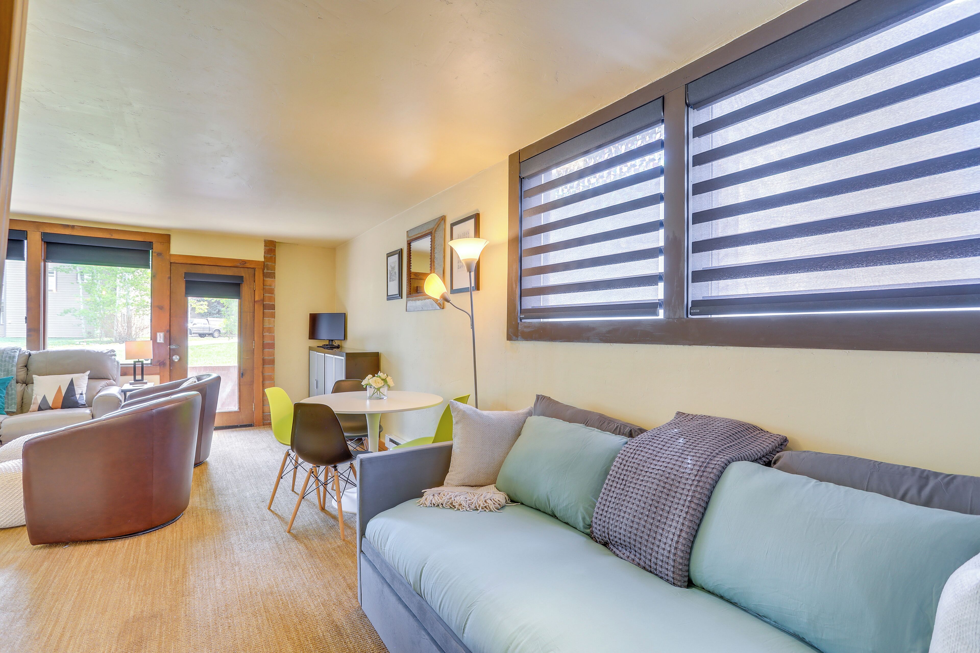 A modern living room with a sofa, chairs, a dining table, and a large window with blinds.