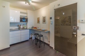 Une cuisine moderne avec des meubles blancs, un four à micro-ondes et un évier. On y trouve deux tabourets et un petit îlot central doté d’une planche à découper et d’une bouilloire.