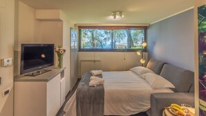 Ein Schlafzimmer mit Bett, Fernseher, Fenster mit Blick auf Bäume und einem kleinen Tisch mit Obst.