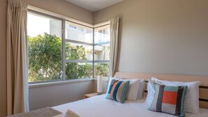A hotel room with a bed, pillows, a window with curtains, and a view of greenery outside. - The Sands - Apartment 23 - Onetangi Beach (Waiheke Island)