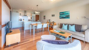 A modern living room with a sofa, ottoman, and coffee table, a kitchen in the background, and a dining area with chairs and a table. - The Sands - Apartment 23 - Onetangi Beach (Waiheke Island)