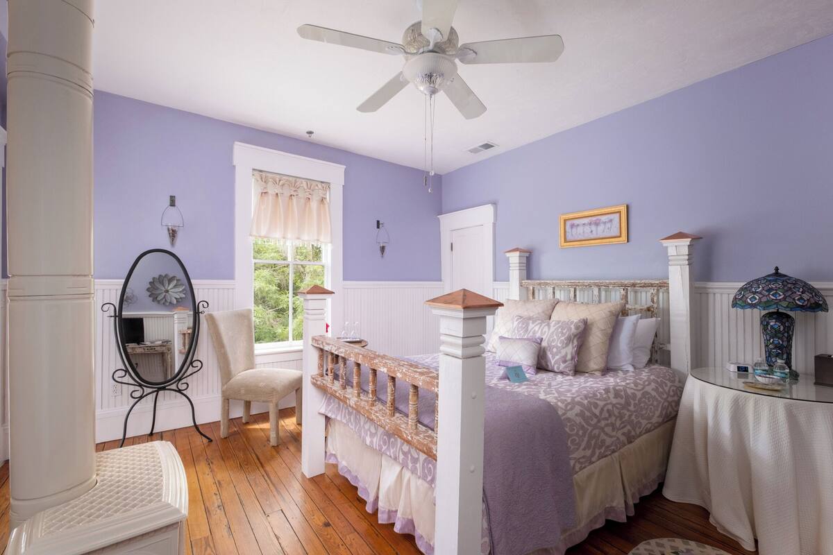 A bedroom with a bed, a chair, a vanity, a ceiling fan, and a window with curtains.