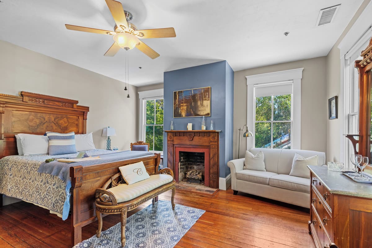 A bedroom with a bed, a wooden headboard, a nightstand, a bench, a fireplace, a sofa, and a dresser.