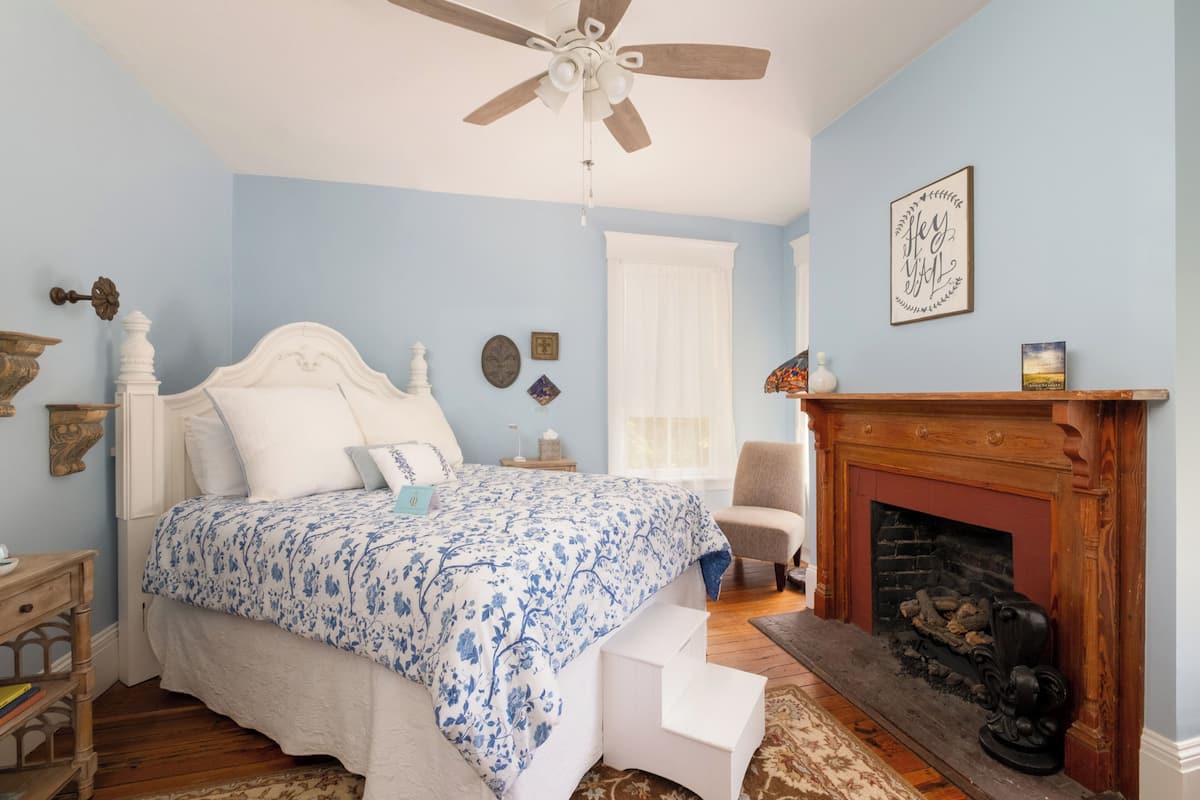 A bedroom with a bed, a fireplace, a ceiling fan, and a window with curtains.
