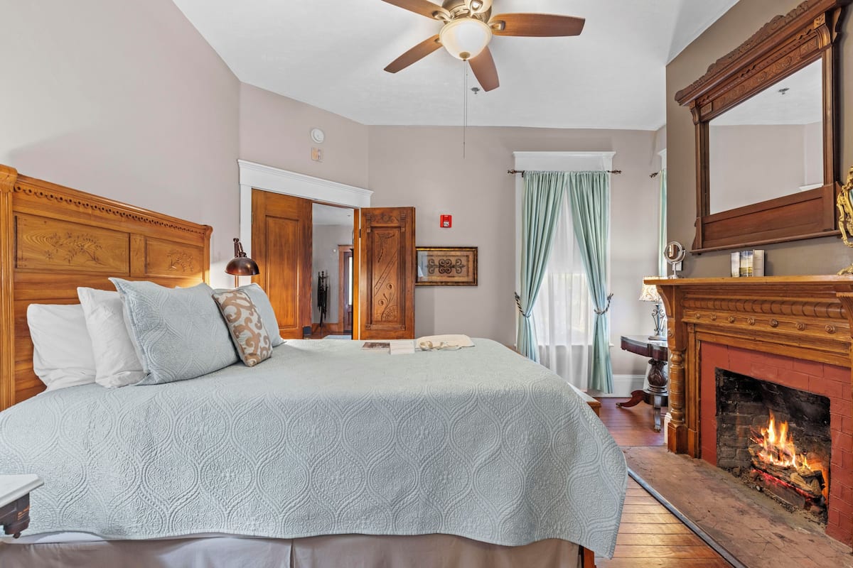A bedroom with a large bed, a wooden headboard, a fireplace, and a ceiling fan.