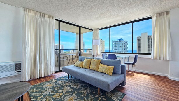 Een moderne hotelkamer met een grijze bank, een klein tafeltje, een stoel, een groot raam met uitzicht op de stad en een balkon.