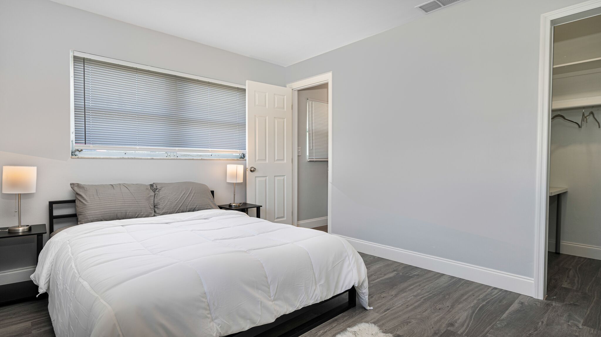 A bedroom with a bed, bedside tables, a window with blinds, a door leading to a closet, and a wooden floor.