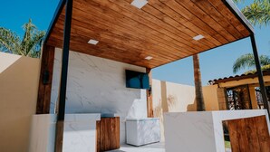 Uma cozinha ao ar livre moderna com bancadas de mármore, vigas de madeira e uma televisão fixada na parede.