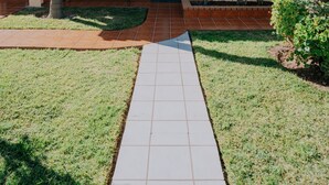 Uma casa com um caminho de ladrilhos que leva à entrada, rodeada por palmeiras e um gramado bem cuidado.