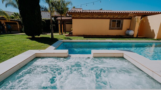 Uma piscina com hidromassagem no centro, rodeada por um gramado bem cuidado e uma casa com telhado em azulejo.