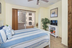 Une chambre à coucher avec un lit, une télévision, une plante et un balcon donnant sur un immeuble.