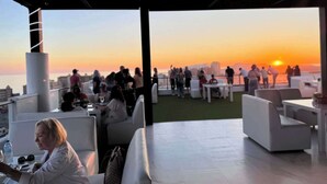 Área externa com mobiliário branco e vista para o pôr do sol sobre o oceano.