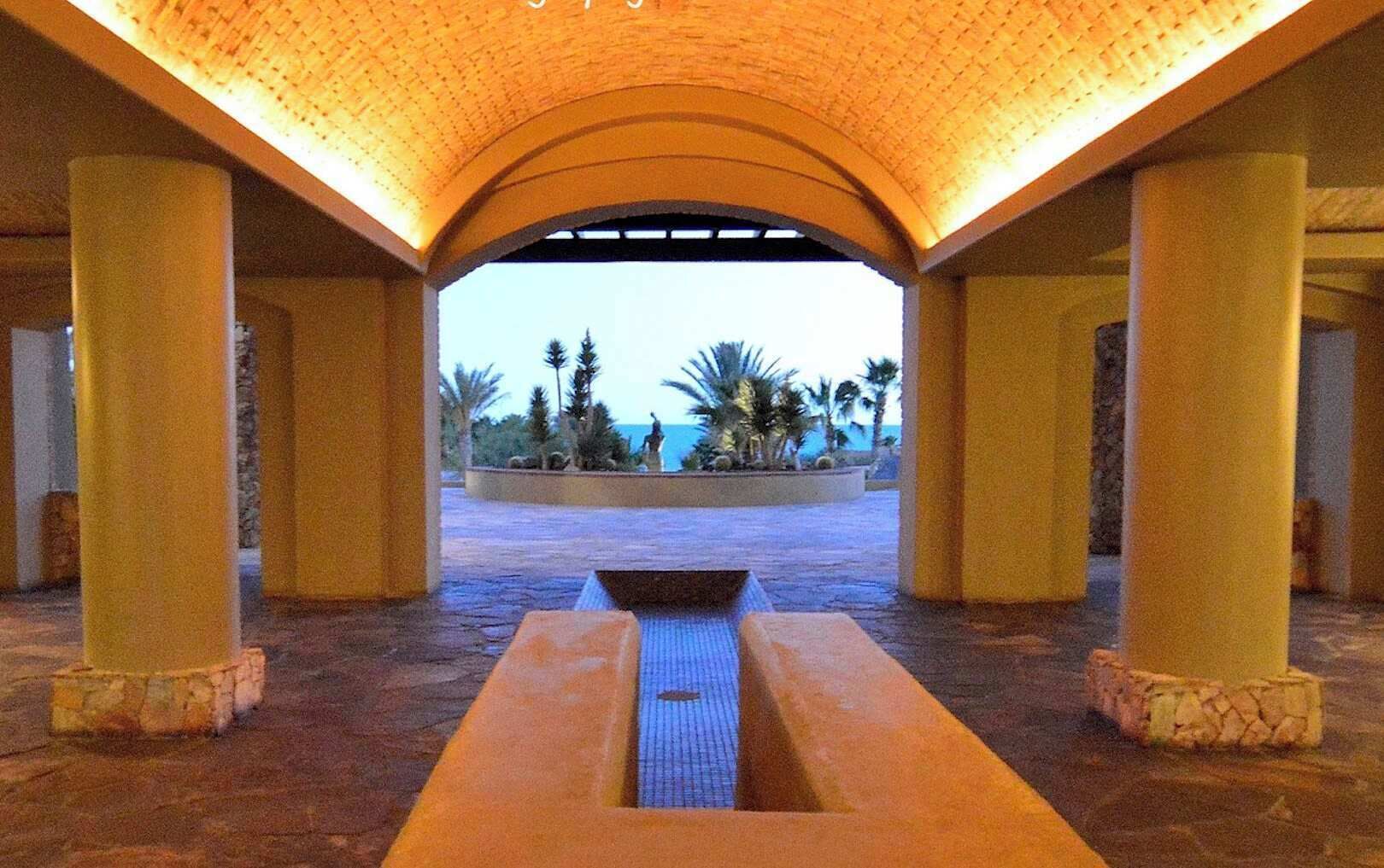 Un espace intérieur doté d’un élément aquatique central, de colonnes et offrant une vue sur des palmiers et l’océan.