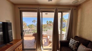A living room with a TV, a sofa, and a balcony with a view of palm trees and buildings. - Fun Sandy Beach Condo B204 at Casa Blanca (Puerto Peñasco)