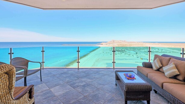 Un balcón con vistas al mar, mobiliario de mimbre y baranda de cristal.