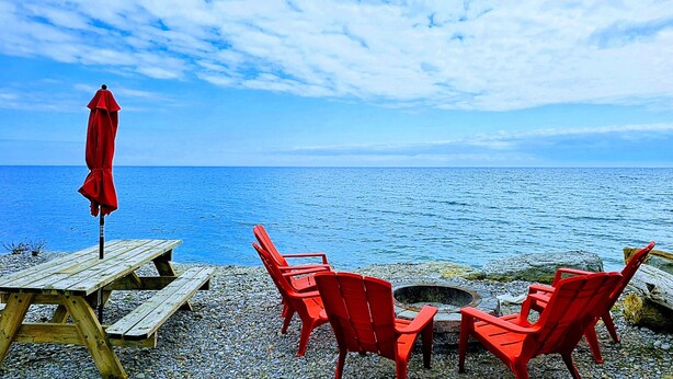 A wooden deck with a picnic table and red chairs, overlooking a calm sea. - Lakeside Dreams: Retreat with Stunning Views, private Beach Access (Bowmanville)