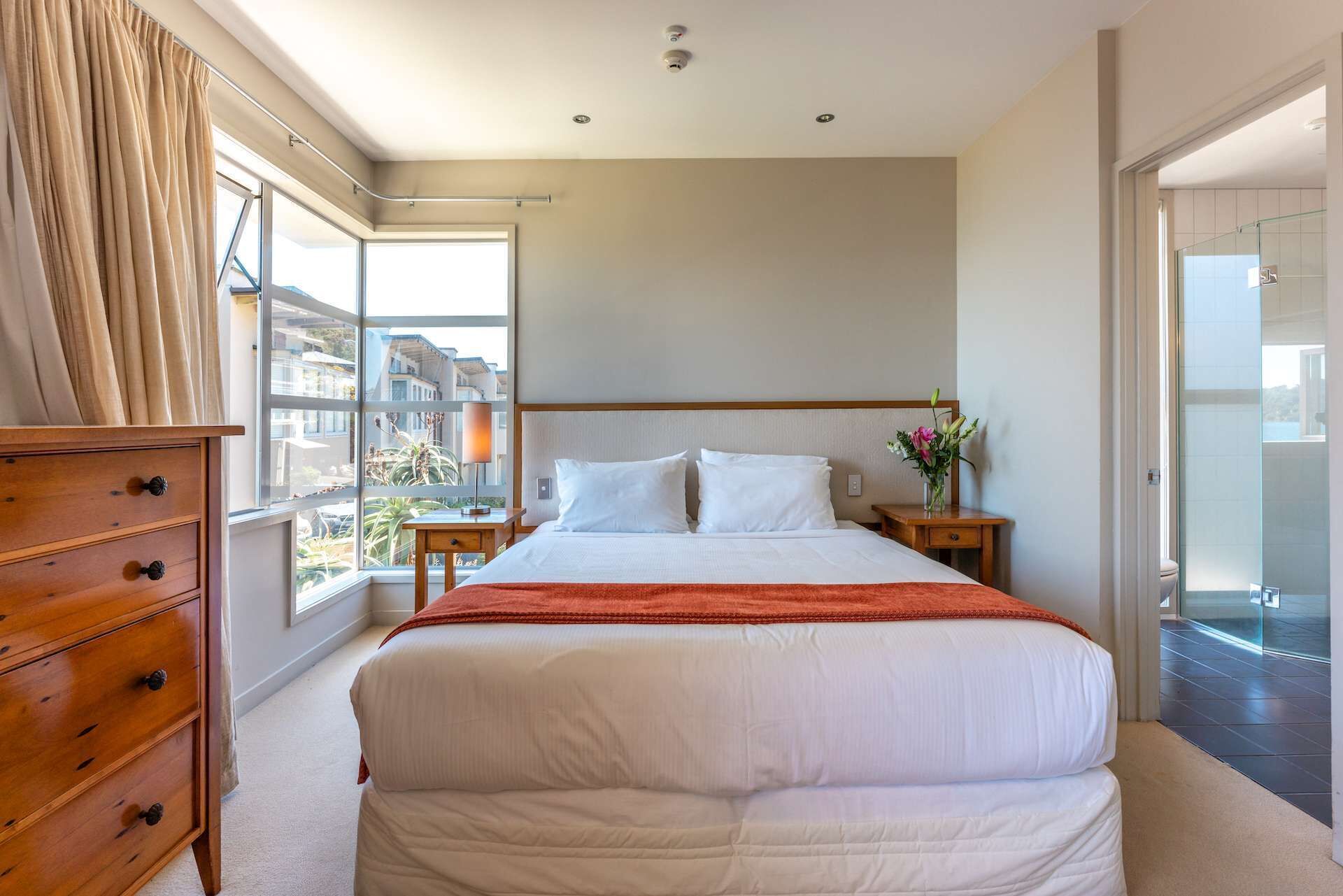 Ein Hotelzimmer mit einem großen Bett, einem Holzschrank, einem Nachttisch mit Lampe und Blick auf die Stadt durch große Fenster.