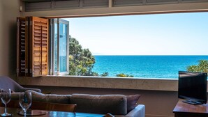 Een kamer met een groot raam met uitzicht op de kust, een bank, een tafel met glazen en een televisie.