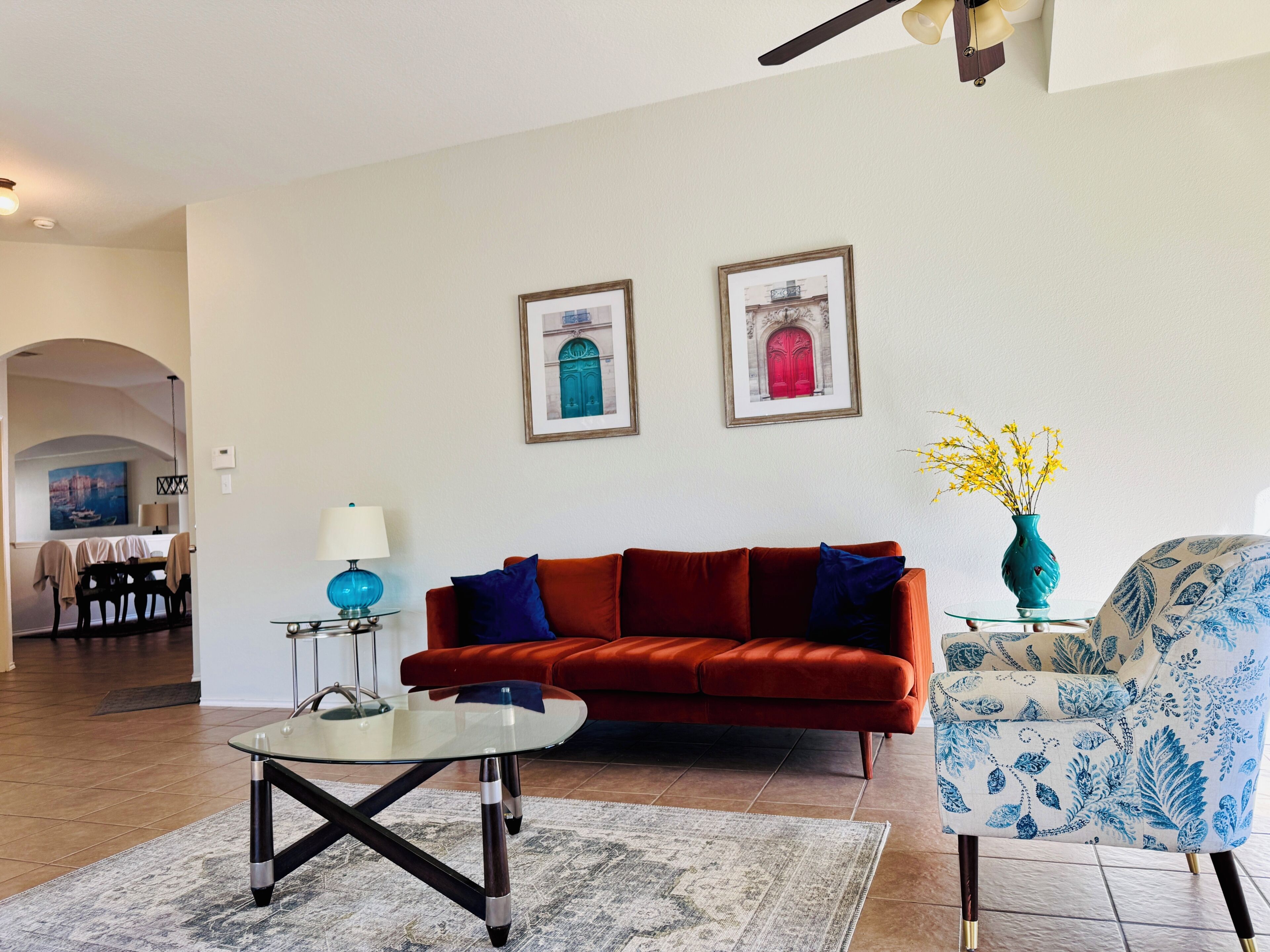 Un salon avec un canapé rouge, une table basse en verre et un fauteuil à motifs. Deux tableaux encadrés sont accrochés au mur, une lampe se trouve sur une table d’appoint et un vase rempli de fleurs est posé sur une petite table.