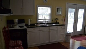 A compact kitchen with white cabinets, a stove, a sink, and a window with blinds. - LYC Cottage #1 (Port Austin)
