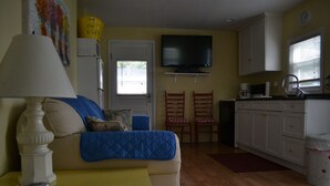 A living room with a sofa, a lamp, a television, and a kitchen area with cabinets and a sink. - LYC Cottage #1 (Port Austin)