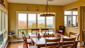 A dining room with a large table, chairs, a chandelier, and a view of autumn foliage through large windows. - Luxury Mountaintop Estate – Spacious Retreat with Breathtaking Views (Bovina Center)