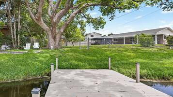 A wooden dock leading to a calm water body, surrounded by lush greenery and a residential area with houses and a fenced property.