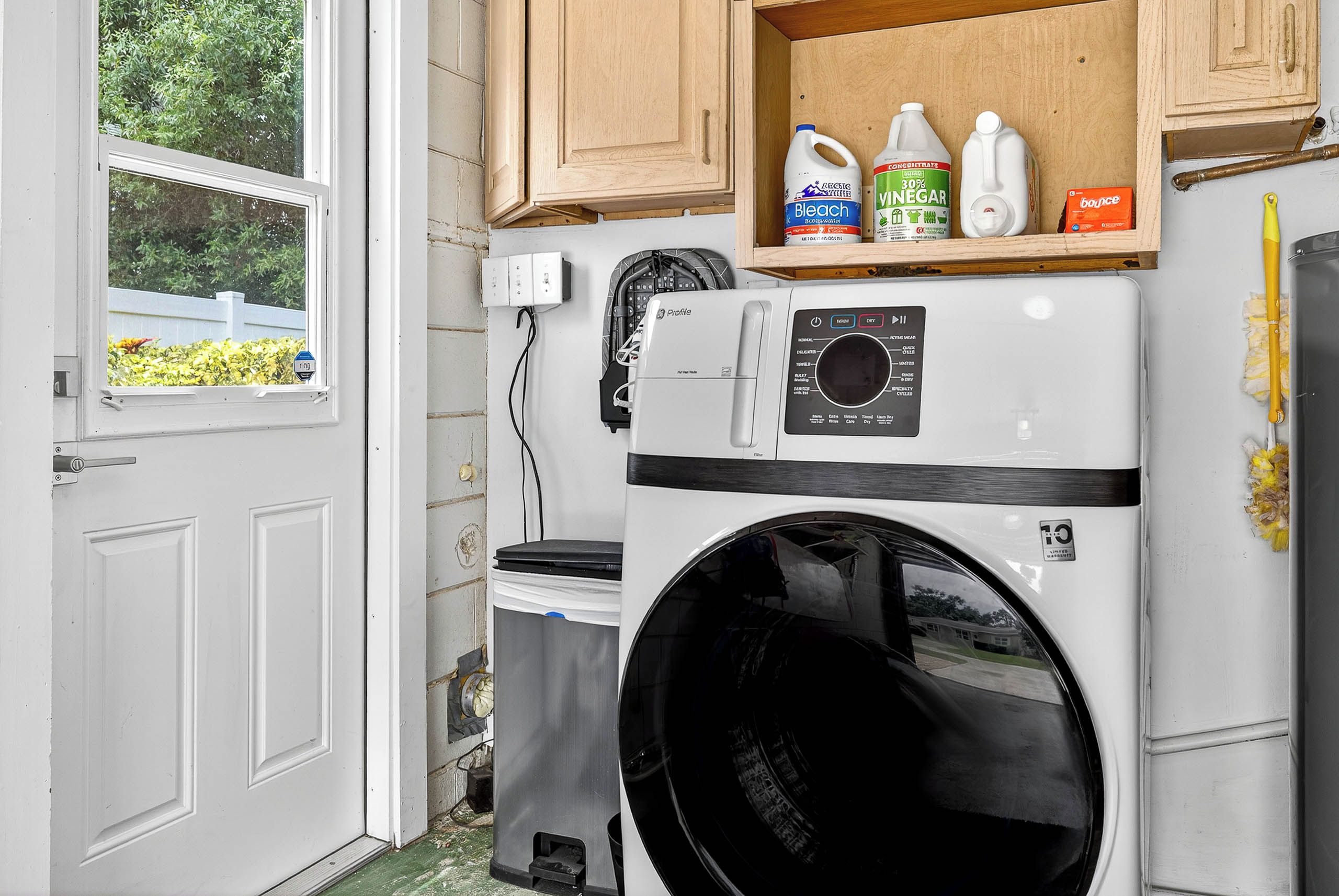 A compact laundry room with a front-loading washer and dryer, a shelf with cleaning supplies, and a window with a view of greenery.