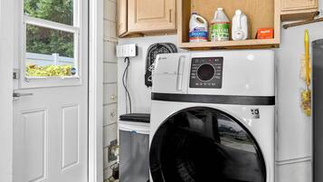 A compact laundry room with a front-loading washer and dryer, a shelf with cleaning supplies, and a window with a view of greenery.